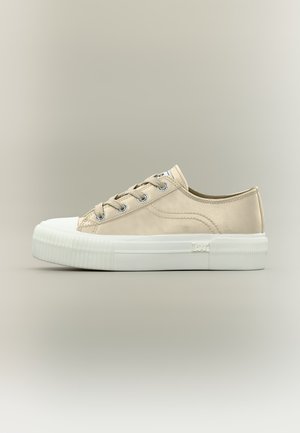Lage beige canvas sneaker met witte rubberen neuskap, dikke witte zool, metalen veterogen en merkdetail aan de zijkant.