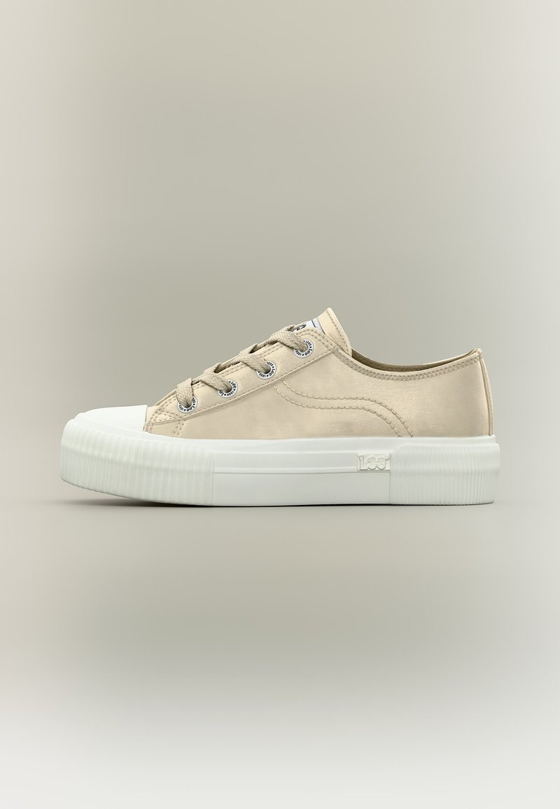 Lavt beige lerretssneaker med hvit gummikappe foran, tykk hvit såle, metallhull og merke på siden.