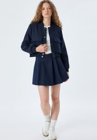 Dunkelblaues Jacket mit zwei Fronttaschen, strukturiertem Material und gerippten Bündchen, kombiniert mit einem plissierten Rock und weißen Sneakers.