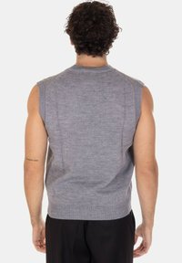 Uomo con capelli ricci che indossa un maglione grigio senza maniche, visto di spalle su uno sfondo bianco.