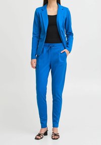 Kvinna iklädd en ljusblå kostym med smala byxor och kavaj, svart linne och svarta öppna klackskor mot en ljus bakgrund.