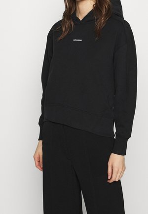 Personne portant un sweat à capuche noir avec le logo « Calvin Klein Jeans » sur la poitrine et un pantalon noir assorti, sur un fond clair uni.