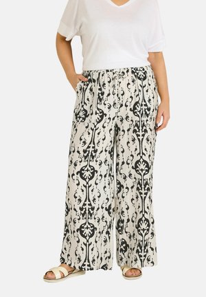 Donna che indossa pantaloni bianchi a gamba larga con motivi floreali neri intricati, abbinati a sandali bianchi e una maglietta bianca a maniche corte.