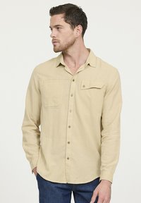 Beige overhemd met lange mouwen en knopen in een lichte stof, met een borstzak en een subtiele textuur, gecombineerd met donkerblauwe jeans.
