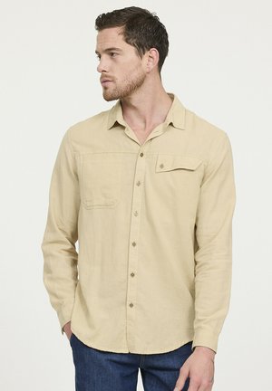 Beige long-sleeve button-up shirt in a lightweight fabric with a chest pocket and subtle texture, paired with dark blue jeans.