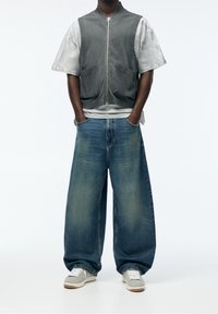 Harmaa liivi, jossa etutaskut, vaaleanharmaan t-paidan päällä; löysät siniset farkut; harmaat ja valkoiset tennarit, joissa on ruskeat yksityiskohdat; minimalistinen muotoilu.