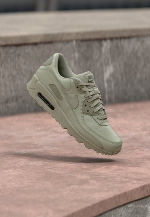 Gaiši bēšas krāsas Nike Air Max sporta kurpes ar redzamu gaisa spilvena daļu, teksturētu ādu un šņorēm, piekarinātas virs akmens virsmas ārā.