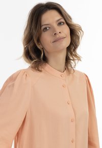 Ljus persikofärgad skjorta med knappar i mandarinstil, sammetslena axlar och en slät textur. Har flera knappar fram.