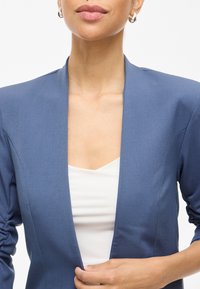 Modrý blazer se štíhlým střihem, s šálovým límcem a nařasenými rukávy, nošený přes bílý top. Hladká látka s matným povrchem.