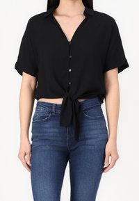 Schwarzes kurzärmliges Hemd mit Knopfleiste und V-Ausschnitt, mit einem gebundenen Vorderteil. Kombiniert mit enganliegenden blauen Jeans. Glatter Stoff, lässige Passform.