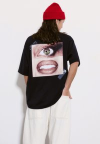 Musta ylisuuria t-paita, jossa on valokuvaprintti silmästä ja suusta, siniset teippikorostukset. Yhdistetty valkoisiin leveälahkeisiin housuihin.