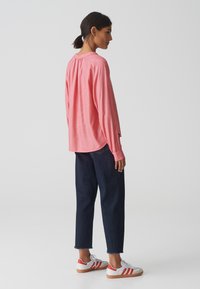 Chemise corail à manches longues avec détail froncé au niveau du cou, associée à un pantalon en denim foncé coupé et des baskets blanches avec des rayures rouges.
