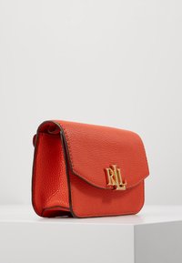 Pomarańczowa skórzana torebka o teksturowanej powierzchni, z logo z złotego metalu z przodu oraz klapką zapinaną na zatrzask. Kompaktowy, prostokątny kształt.