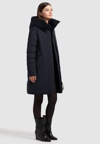 Abrigo acolchado azul marino con capucha, con corte recto, bolsillos laterales y un acabado suave y mate. Lleva puesto sobre un vestido oscuro y botas de cuña negras.