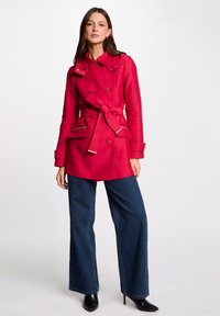 Cappotto rosso impermeabile con vita accentuata da una cintura, chiusura a bottoni e tasche frontali doppie. Abbinato a jeans a gamba larga e tacchi a punta neri.