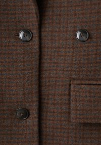 Ruskea ruudullinen takkutekokangas, jossa on tumma sininen viivoitus, kaksi mustaa nappia ja sivutasku.