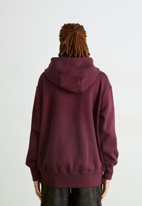 Moletom oversized bordô com capuz grande, punhos e barra canelados; material de algodão macio; vista traseira mostrando cabelo trançado.