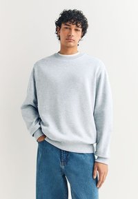 Felpa grigio chiaro con collo tondo, polsini e orlo a coste, abbinata a jeans di denim blu. Il tessuto sembra morbido e leggermente strutturato.