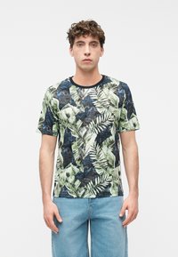 Νεαρός άνδρας όρθιος φορώντας μπλουζάκι με σχέδιο τροπικά φύλλα και ανοιχτό μπλε τζιν μπροστά σε απλό λευκό φόντο.