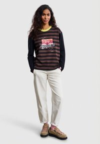 Žena s dugom tamnom kosom nosi crno-smeđu prugastu majicu dugih rukava s natpisom "Hogwarts", bijele hlače i smeđe planinarske cipele.