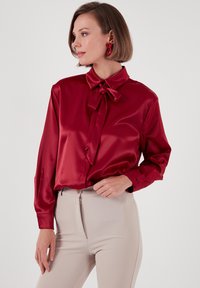 Red satin blouse with a high collar and bow tie at the neck. Long sleeves with button closures. Paired with beige fitted pants.