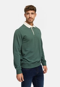 Groene polo met lange mouwen en een crèmekleurige kraag, gemaakt van zacht materiaal. Heeft een knoopsluiting met twee knoppen en ribgebreide manchetten.