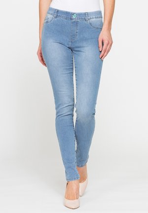 Donna che indossa jeans skinny azzurri chiari e tacchi alti beige, in piedi con una gamba leggermente avanti su uno sfondo bianco uniforme.