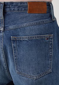 Plave traperice s kožnom oznakom Tommy Hilfiger, vidljivim šavovima na stražnjim džepovima i klasičnim ravnim krojem.