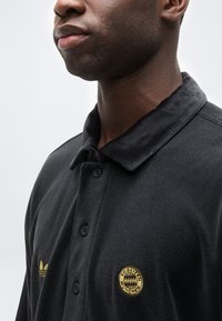 Κοντινό πλάνο ενός άνδρα που φοράει μαύρο πόλο μπλουζάκι με λογότυπα της FC Bayern Munich και της Adidas σε κίτρινο στο στήθος.