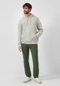 Mężczyzna stoi ubrany w strukturalny jasnoszary hoodie, zielone spodnie i białe tenisówki na jednolitym jasnym tle.