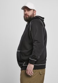 Felpa nera con cuciture grigie, tasca frontale a marsupio, cappuccio regolabile con cordino, indossata con pantaloni khaki, vista di profilo.