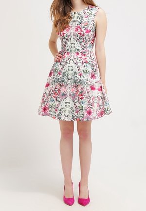 Woman wearing sleeveless white dress with pink floral pattern and pink high heels, standing with one hand on hip against plain background.