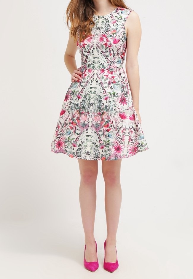 Femme portant une robe blanche sans manches avec un motif floral rose et des talons hauts roses, debout avec une main sur la hanche sur un fond uni.