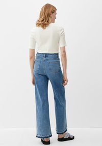Jean en denim bleu à jambe large et taille haute, avec un ourlet effiloché et des poches arrière minimales, associé à un haut ajusté à manches courtes blanc.