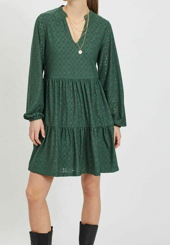 Grön lagd klänning med långa ärmar, ögonhålsmönster, V-ringning och en avslappnad passform. Bärs tillsammans med lager av halsband och knähöga svarta stövlar.