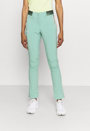 Personne portant un pantalon ajusté vert clair avec des poches zippées et des baskets blanches, debout devant un fond blanc uni.