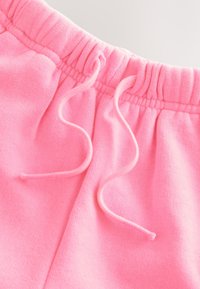 Gros plan de la ceinture d’un pantalon de jogging rose avec bande élastique et cordons lâches sur fond clair.