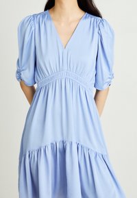 Sininen mekko, joka on valmistettu sileästä kankaasta. Mekossa on V-aukkoinen kaula-aukko, puffi hihat ja kootun vyötärön lisäksi kerroksellinen, virtaava helma.