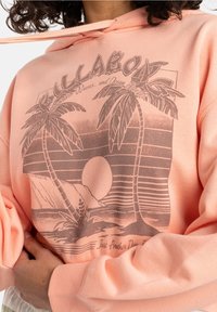 Sudadera rosa con capucha que presenta un gráfico de una palmera, un atardecer y el texto "Solo otro día". Textura suave, corte holgado, diseño informal.