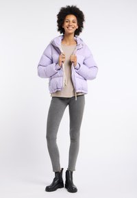 Lila pufferjacka med dragkedja fram och ståkrage, ihop med en beige topp och grå skinny jeans, bärandes svarta ankboots.