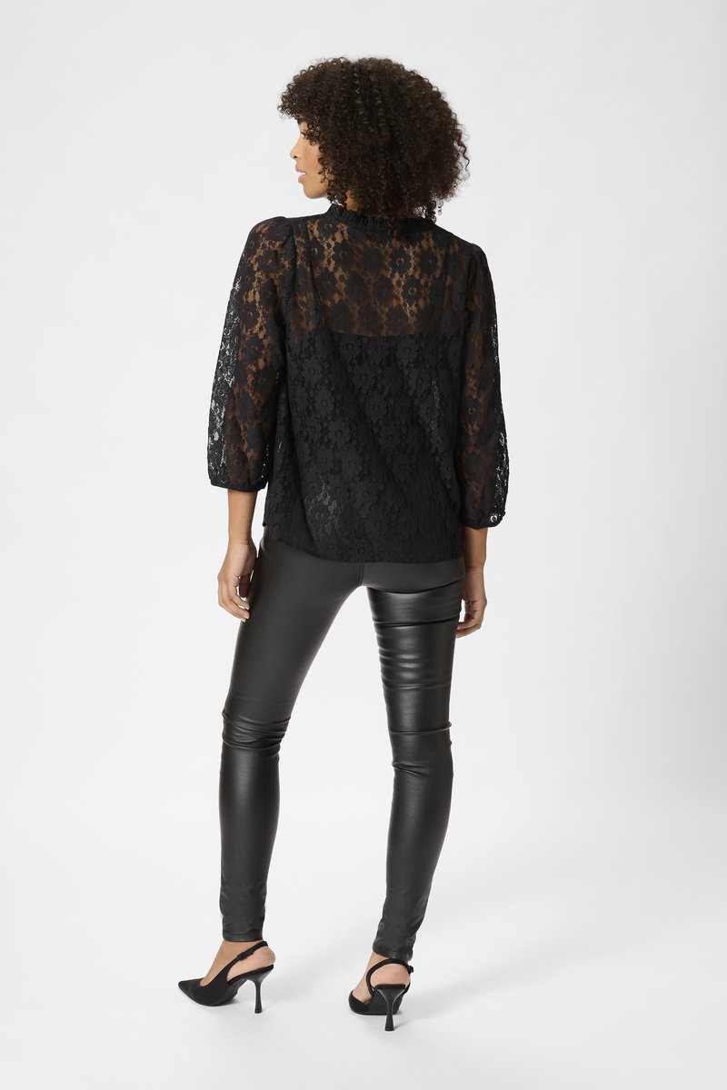 Top de encaje negro con mangas largas, que presenta un escote con volantes y un patrón floral. Combinado con leggins de cuero negro y tacones.