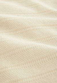 Tissu textile dans une teinte beige clair avec un motif texturé de petits carrés et de lignes horizontales, montrant une texture douce et lisse.