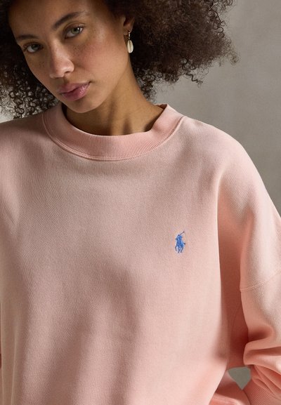 Φούτερ σε ανοιχτό ροζ χρώμα με στρογγυλή λαιμόκοψη, χαλαρή γραμμή στους ώμους και μικρό μπλε λογότυπο στο αριστερό στήθος. Απαλή, υφή υφάσματος με υφή.