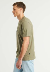 Alyvuogių žalios spalvos trumpas rankovės marškinėliai su apvalia iškirpte, pagaminti iš medvilnės, su šonu etiketė ir paprastu logotipu ant krūtinės. Dėvimos šviesiai mėlynos džinsinės kelnės.