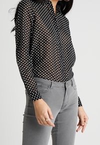 Black sheer blouse with white polka dots, button-down front, and a striped pattern on one side. Paired with gray fitted jeans.