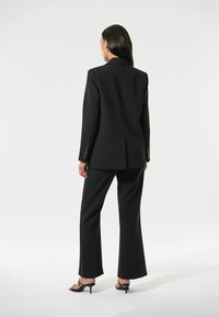 Blazer nero su misura con uno spacco singolo, abbinato a pantaloni coordinati. Tessuto liscio, spalle strutturate e design elegante. Sandali neri con tacco.