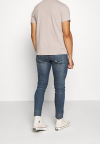 Herrarnas beige t-shirt med kort ärm, åtsittande blå jeans och vita högskoor. Jeansen har lätt blekning och accentuerande sömmar.