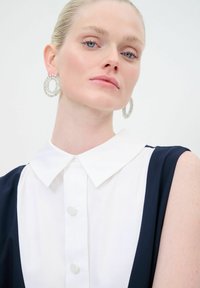 Blusa blanca con cuello, con acentos en azul marino, que presenta un cierre de botones y grandes pendientes de cristal circular, destacando un diseño limpio y moderno.