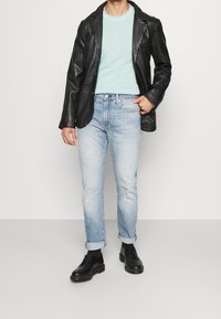 Blazer in pelle nera sopra un maglione verde menta chiaro, abbinato a jeans azzurri chiari con risvolti e scarpe nere stringate. Texture lisce e vestibilità casual.