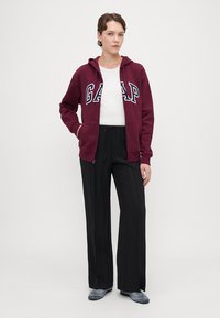 Sudadera burdeos con el logo "GAP", combinada con pantalones anchos negros y zapatos grises. La sudadera cuenta con un zipper frontal y dos bolsillos laterales.